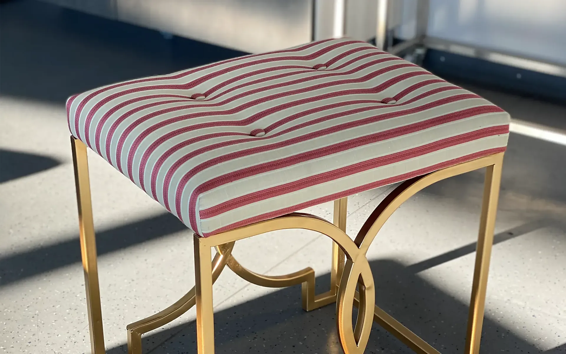 Ein rechteckiger Hocker mit einem rot-weiß gestreiften, gesteppten Kissen auf einem dekorativen, goldfarbenen Metallgestell.