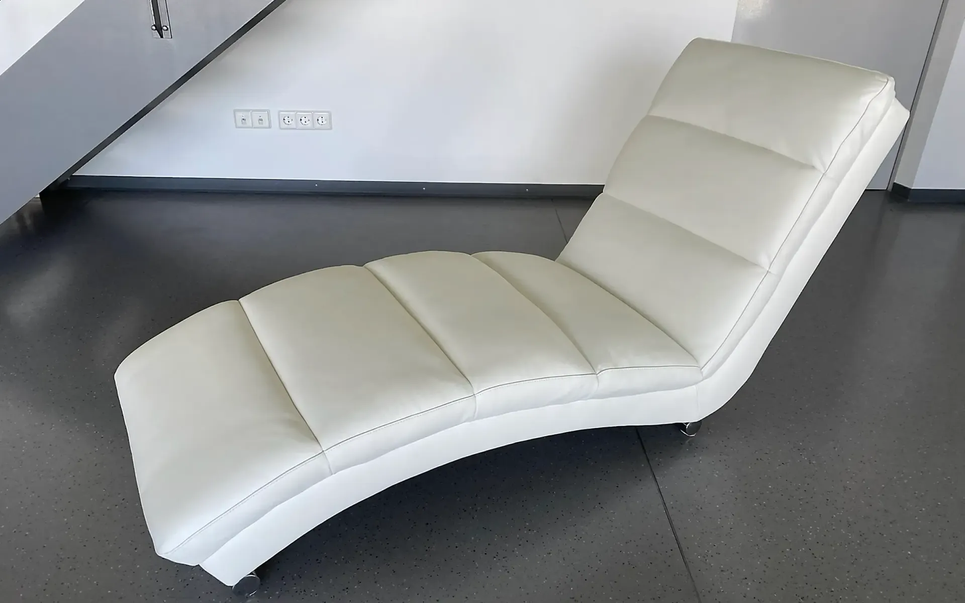 Ein geschwungener, weißer Ledersessel steht auf einem glatten grauen Boden vor einer weißen Wand.