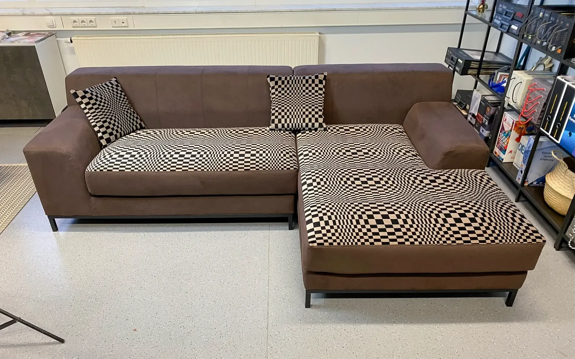 Ein L-förmiges braunes Sofa mit gemusterten Sitzkissen und passenden Zierkissen steht in einem Zimmer mit einem Regal.