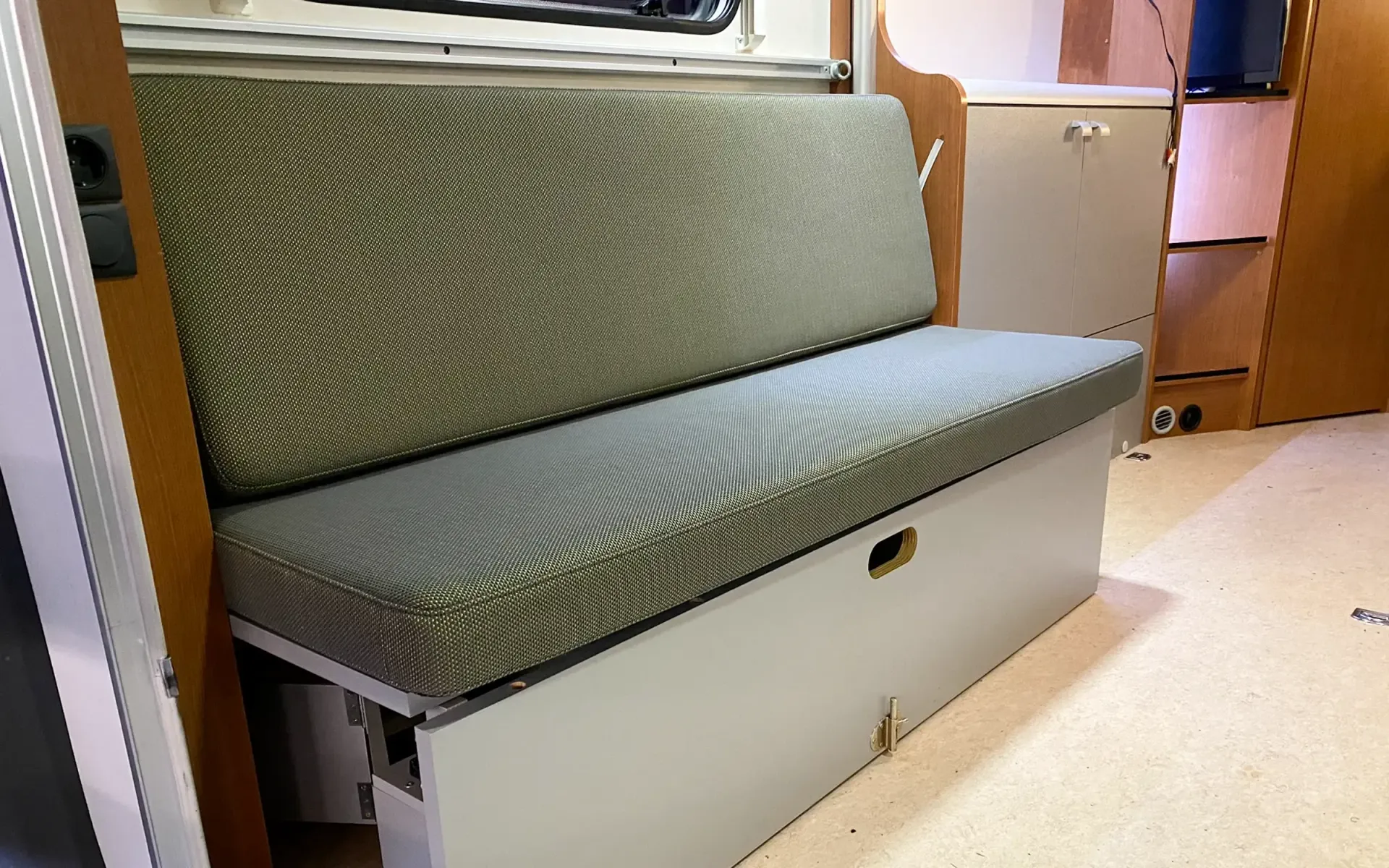 Eine salbeigrüne, gepolsterte Sitzbank mit weißem Staufach im Innenraum eines Wohnmobils.