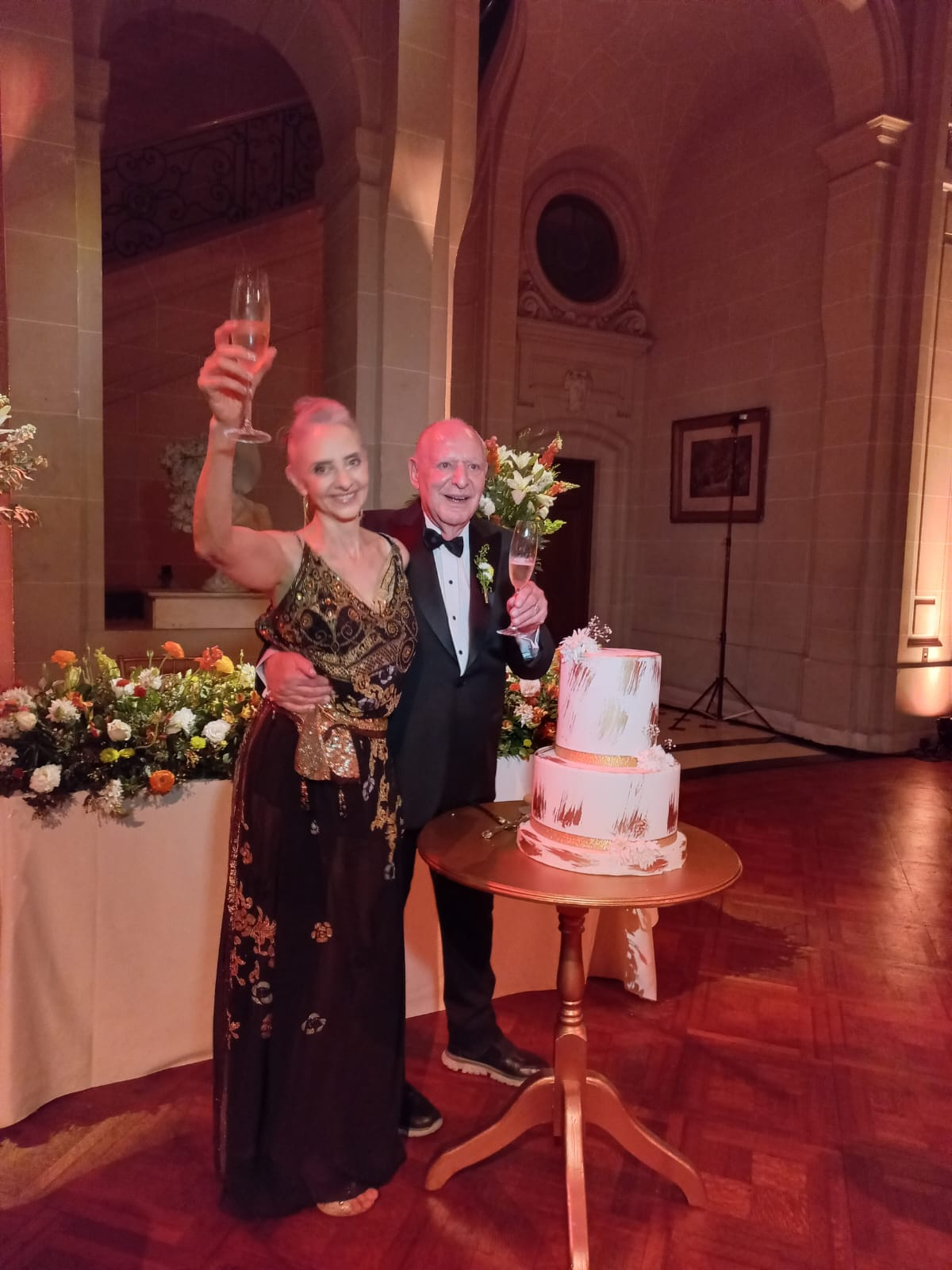 Una pareja vestida de gala brinda con champán junto a un pastel de bodas de varios pisos en una celebración en un gran salón.