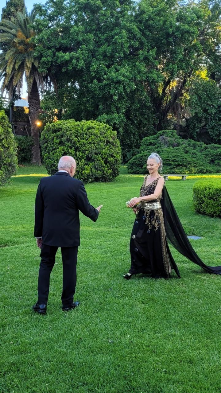 En un jardín exuberante, una persona vestida con un traje negro hace un gesto hacia otra que lleva un vestido negro vaporoso y ornamentado.