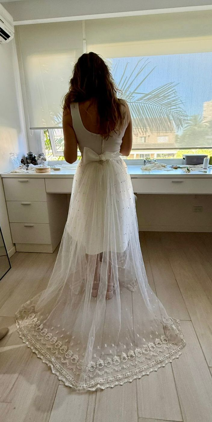 Una persona aparece de pie frente a una ventana, vestida con un vestido blanco que incluye una sobrefalda transparente de encaje bordado y un lazo en la espalda.