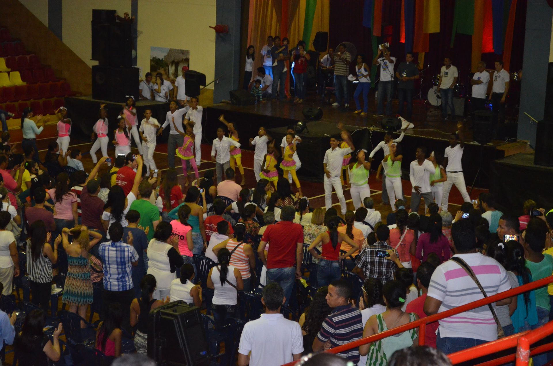 Un grupo de bailarines con trajes blancos y coloridos actúan en un escenario ante un público en un gran recinto cerrado.