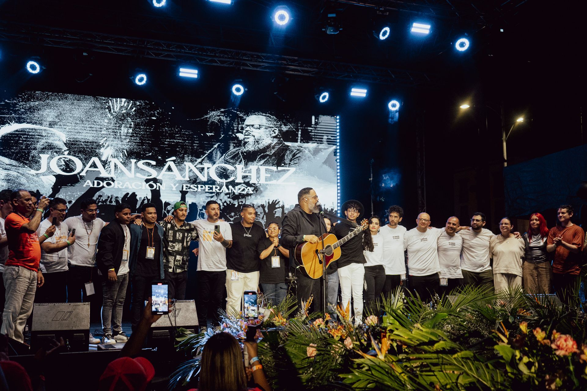 Un numeroso grupo de personas se encuentra de pie sobre un escenario de conciertos brillantemente iluminado