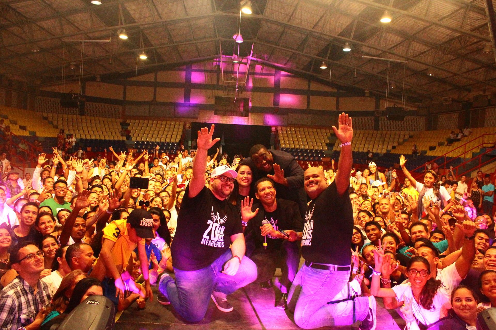 Un grupo de personas con camisetas negras idénticas, arrodilladas en un escenario, saludan a una gran multitud que las aclama en un recinto cubierto.