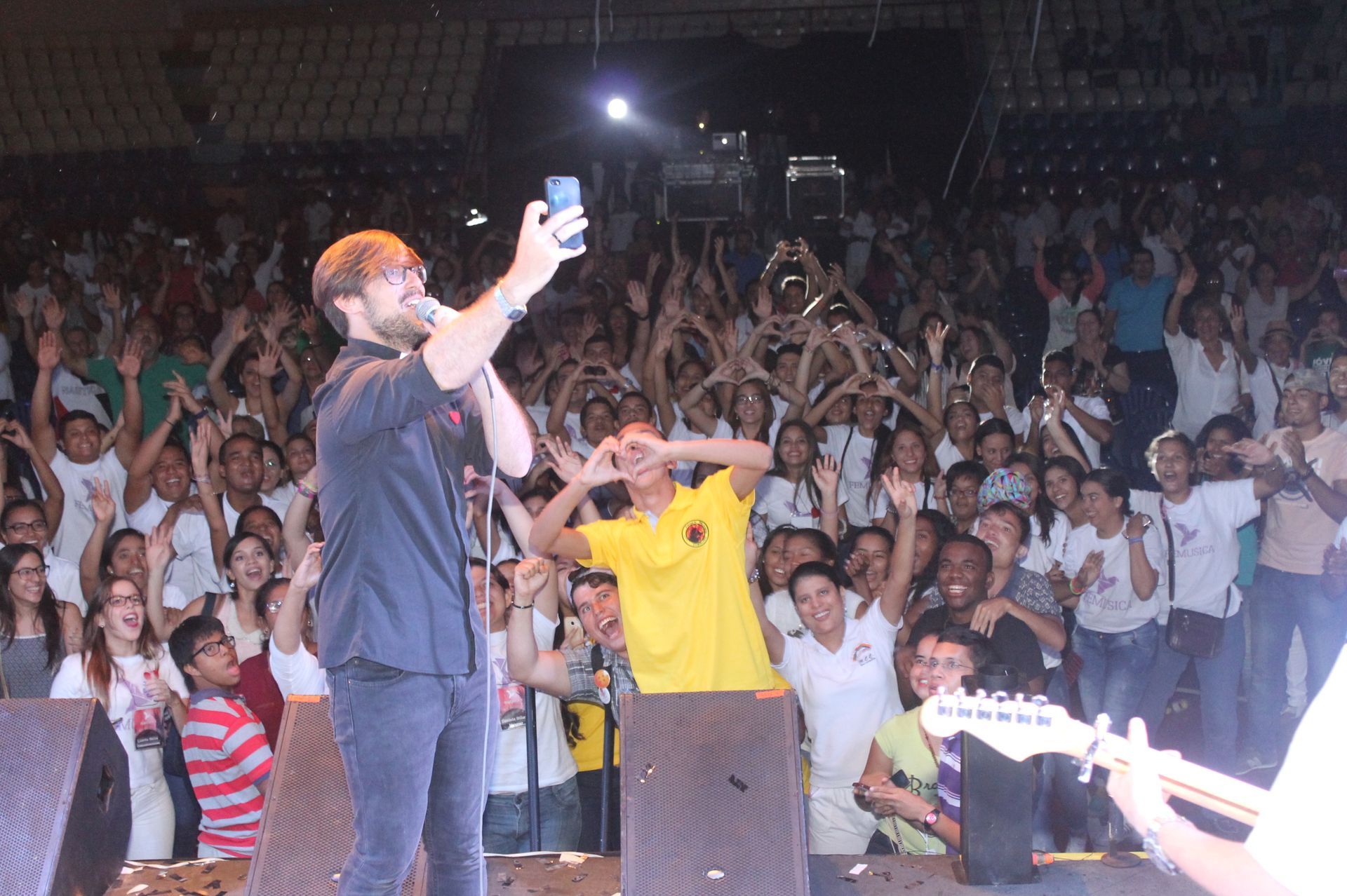 Un artista se toma una selfie en el escenario ante una gran multitud que lo aclama en una sala de conciertos con poca luz.