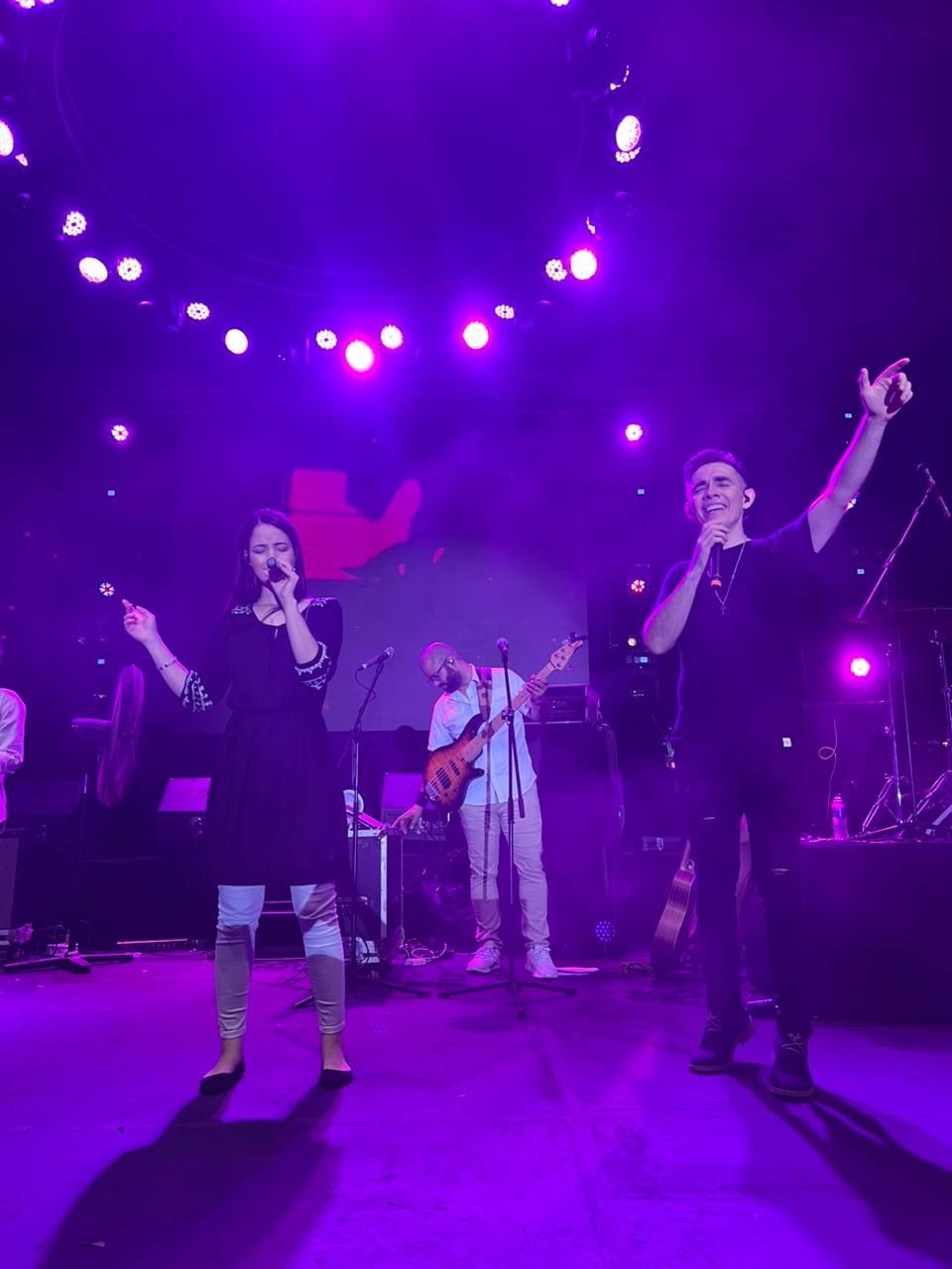Dos cantantes actuando en un escenario con iluminación tenue de color púrpura, acompañados por un guitarrista.