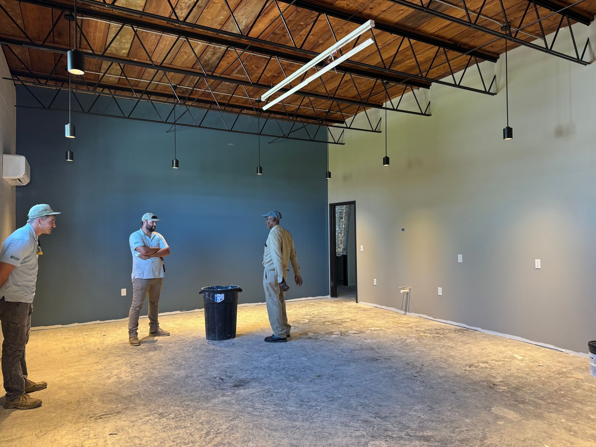 Three men in a mostly empty room with gray walls and a high ceiling are discussing something.