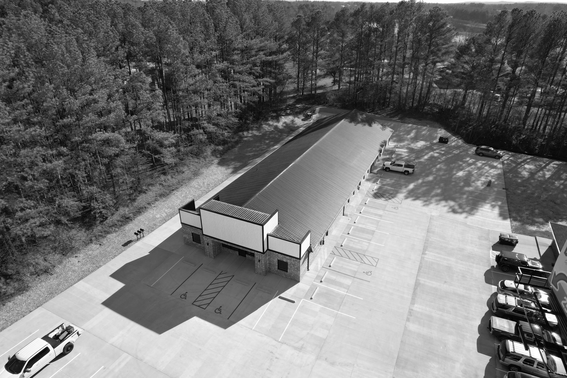 Black and white aerial view of a long building with parking, surrounded by trees.