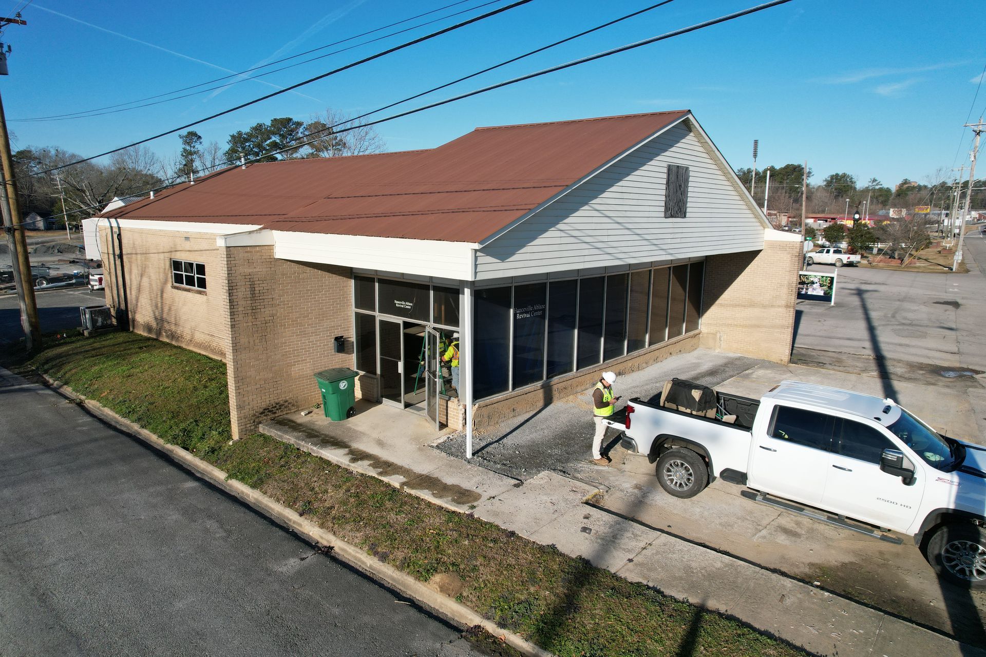 Exterior of a brick building with a brown roof. Two workers near the entrance, a white truck parked on the side.