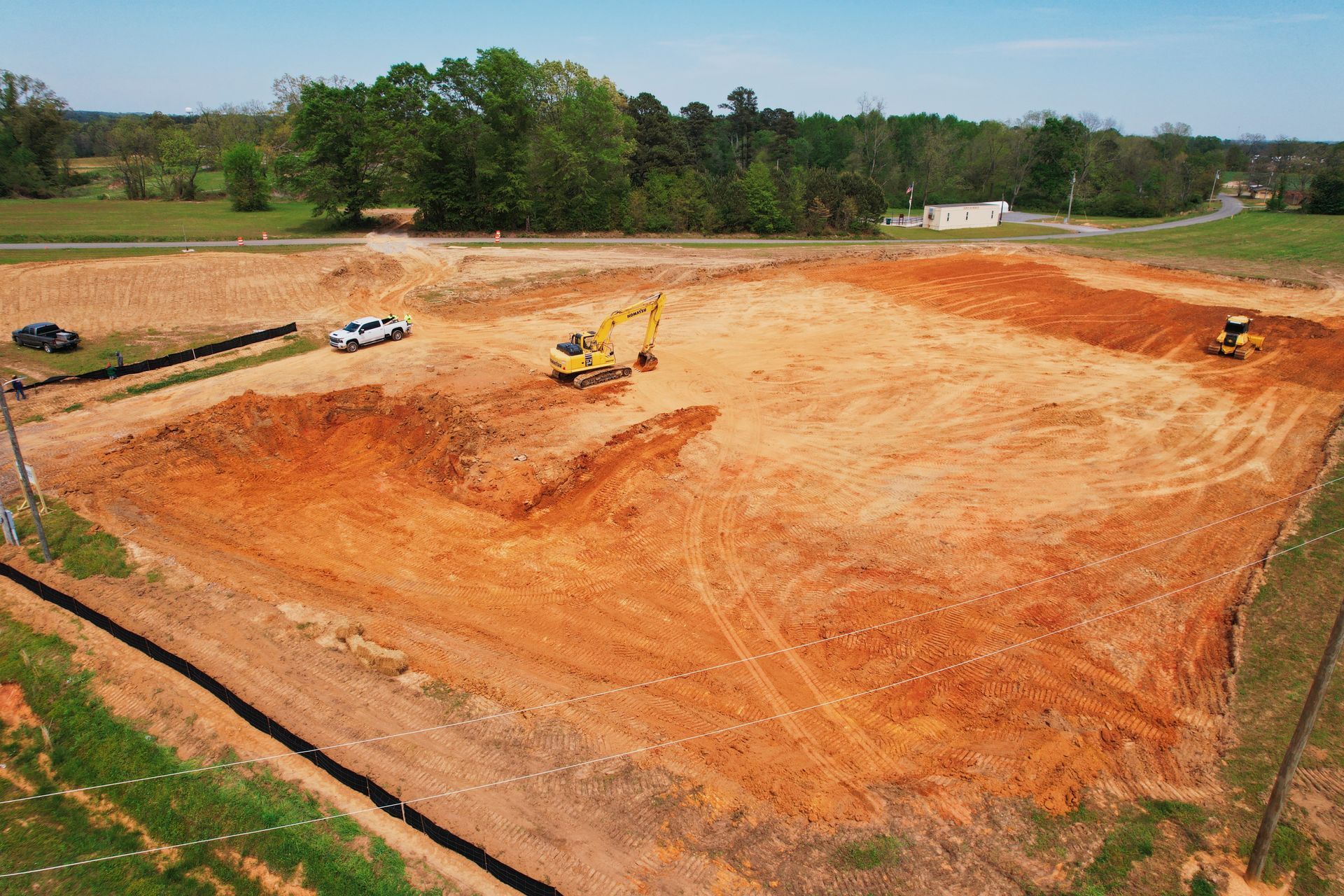 Construction site with heavy machinery grading red dirt. Trees and small building in background.