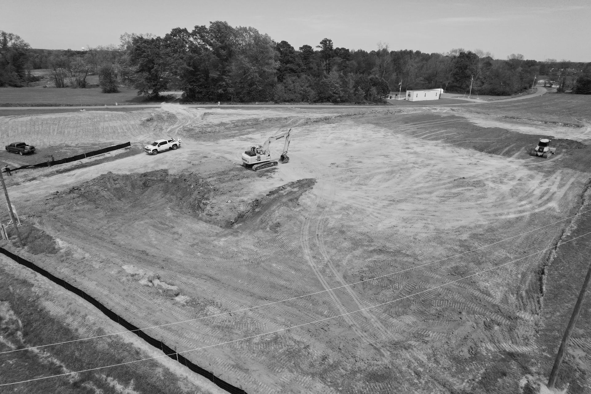 Construction site: dirt mounds, vehicles, trees, and small building in the background.