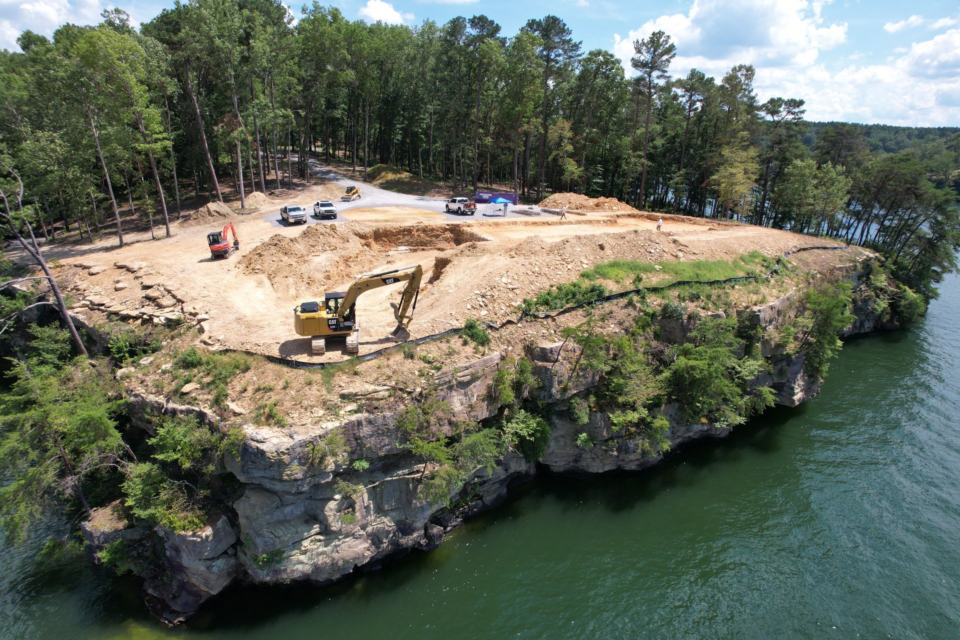 Excavation on a rocky peninsula with machinery, trees, water, and vehicles.