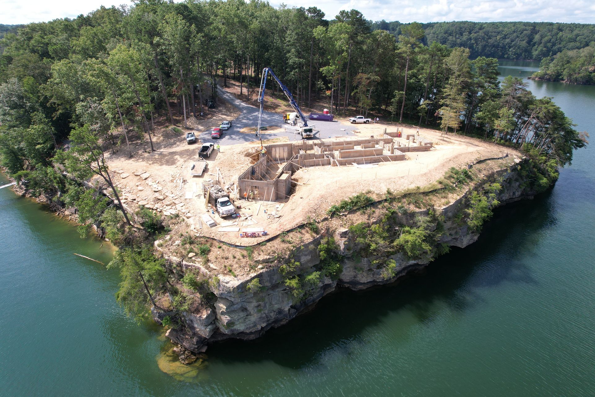 Construction site on a rocky island surrounded by water. Trucks, cranes, and building materials are present. Green trees.