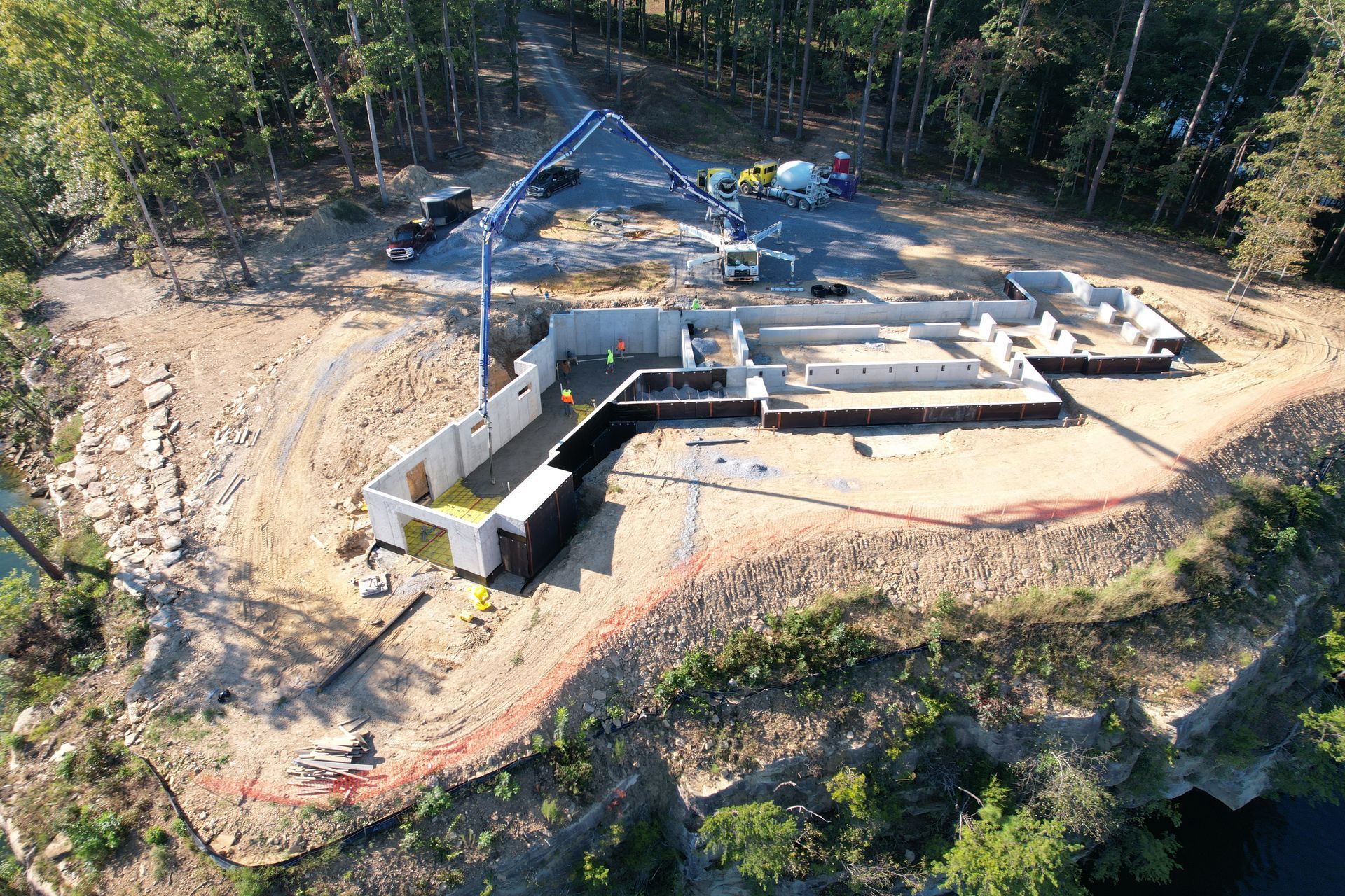 Construction site: foundation poured, concrete pump truck, building on a hill, forest in background.