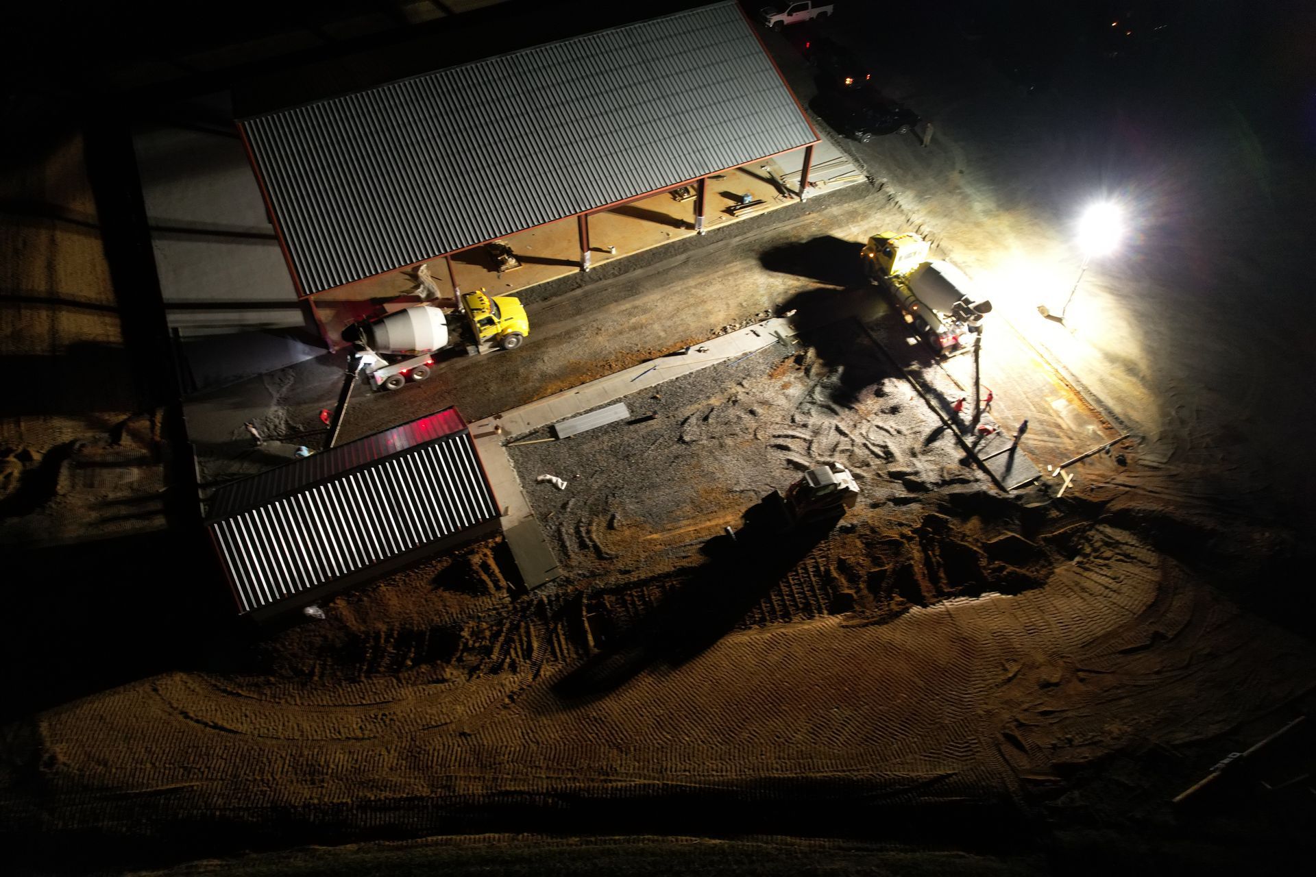 Aerial view of a farm structure at night lit by a spotlight; corrugated metal roof, equipment, and a dirt path.