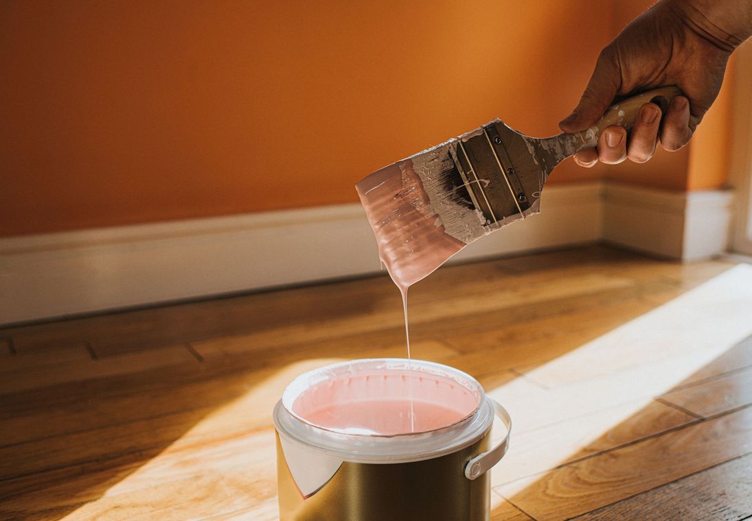 A person holding a paint brush dripping pink paint into a bucket, in a room with wood floor.