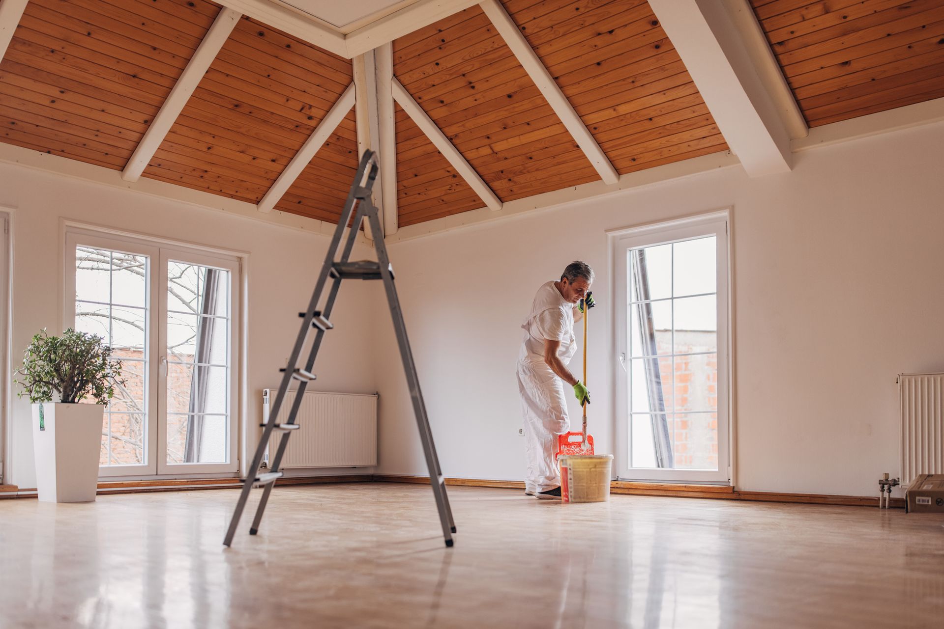 Painter in white overalls standing in an empty room, next to ladder and paint bucket, painting the wall.