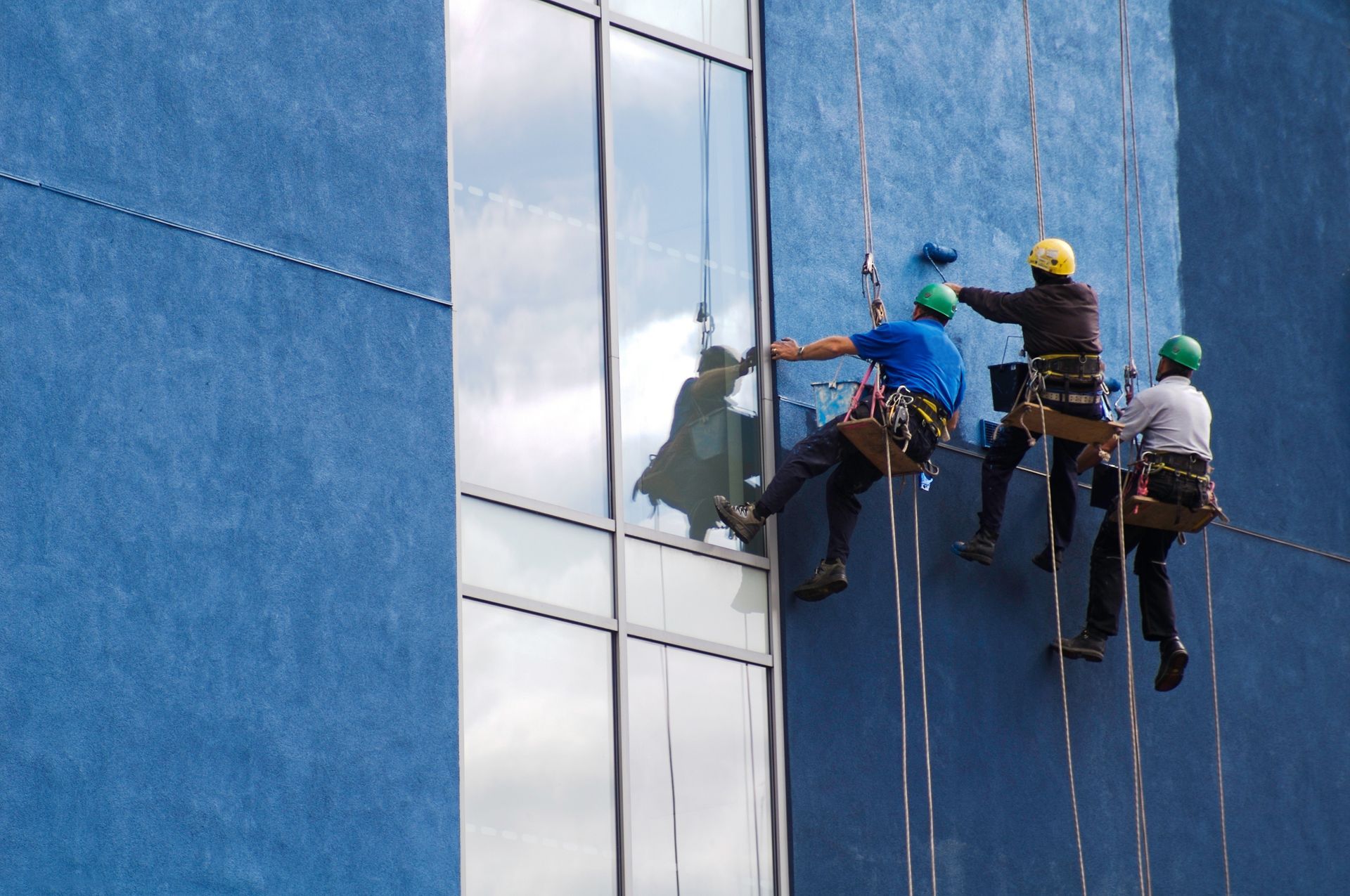 Workers on ropes cleaning windows of a blue building.