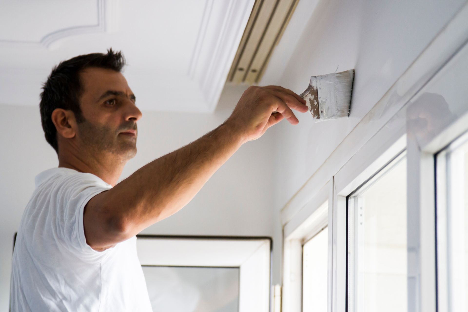 Man in white shirt using a putty knife to apply compound to a wall near a window.