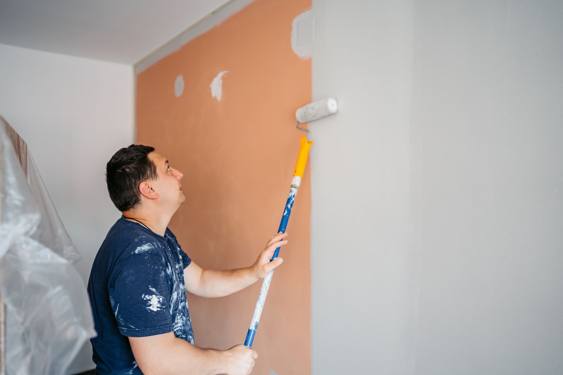 Man painting wall with roller; light-colored paint against peach-colored wall. Indoor setting.