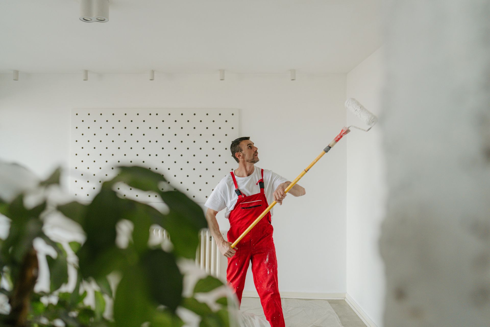 Painter in red overalls, white shirt, using a paint roller on a white wall, inside a room.