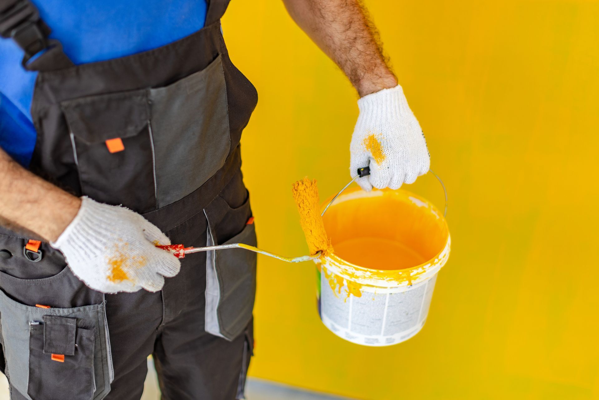 Person in overalls painting a yellow wall with a roller, wearing gloves, holding a bucket of yellow paint.