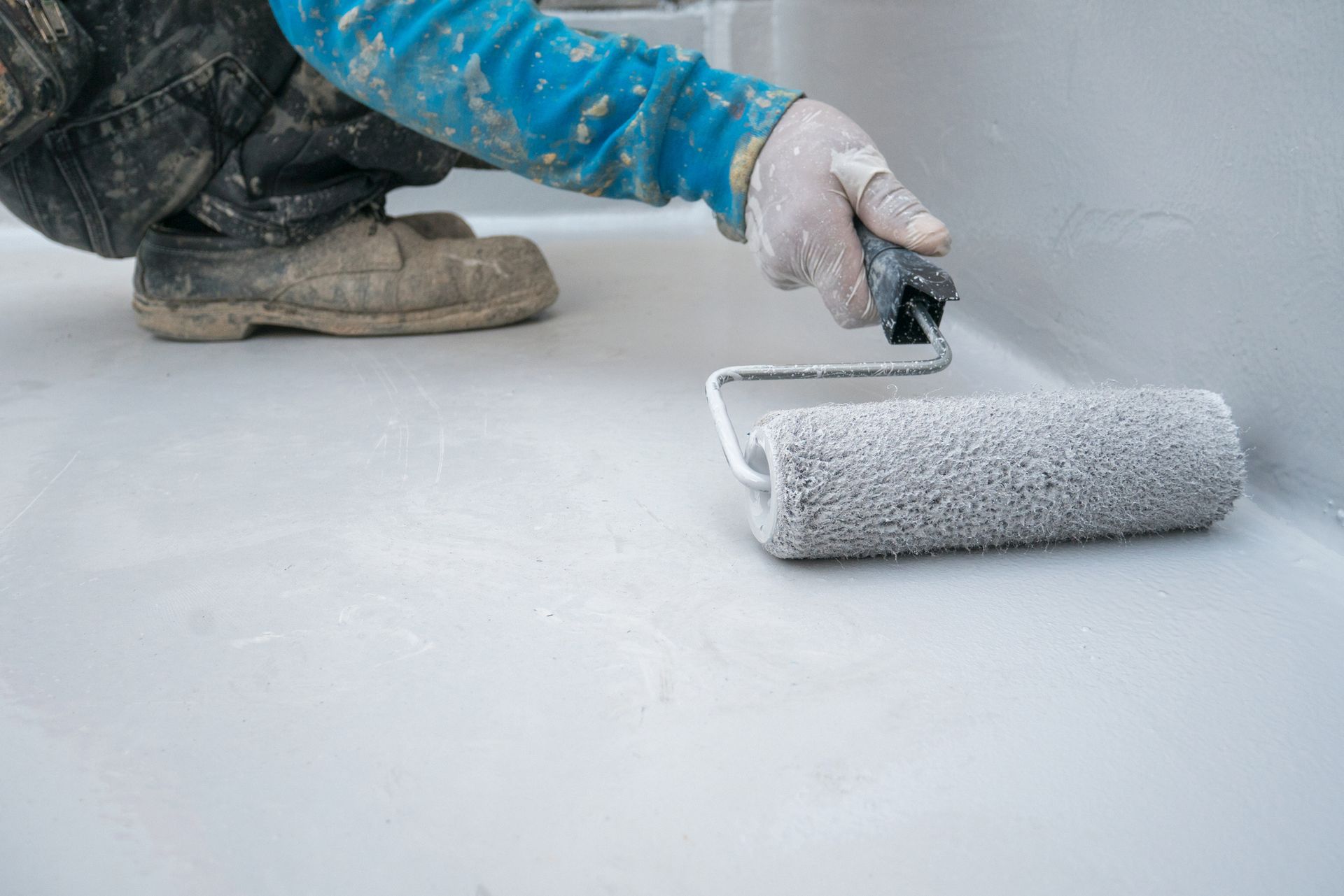 Person in blue shirt and dirty boots painting a light gray surface with a roller.