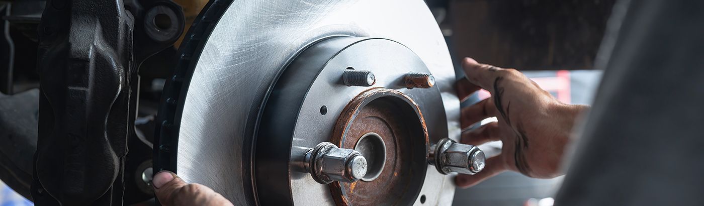 A person's hands holding a new, silver car brake rotor in front of a vehicle's wheel assembly.