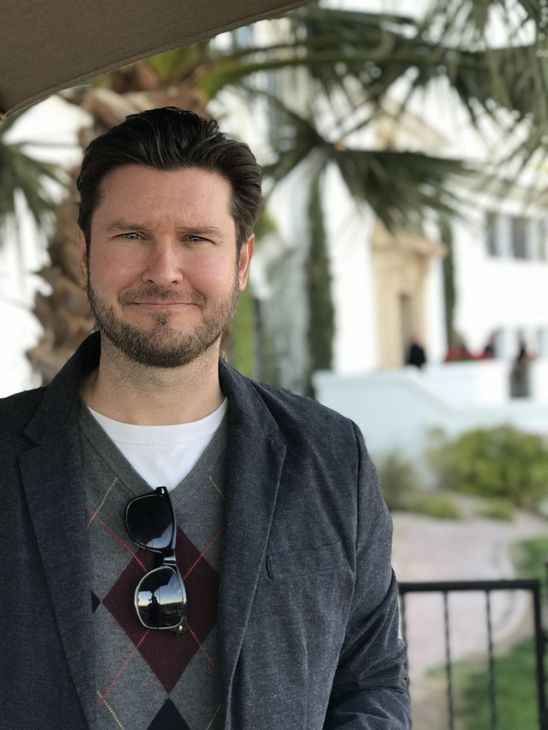 a man with a beard and sunglasses is standing in front of a palm tree. His name is Jesse Clark