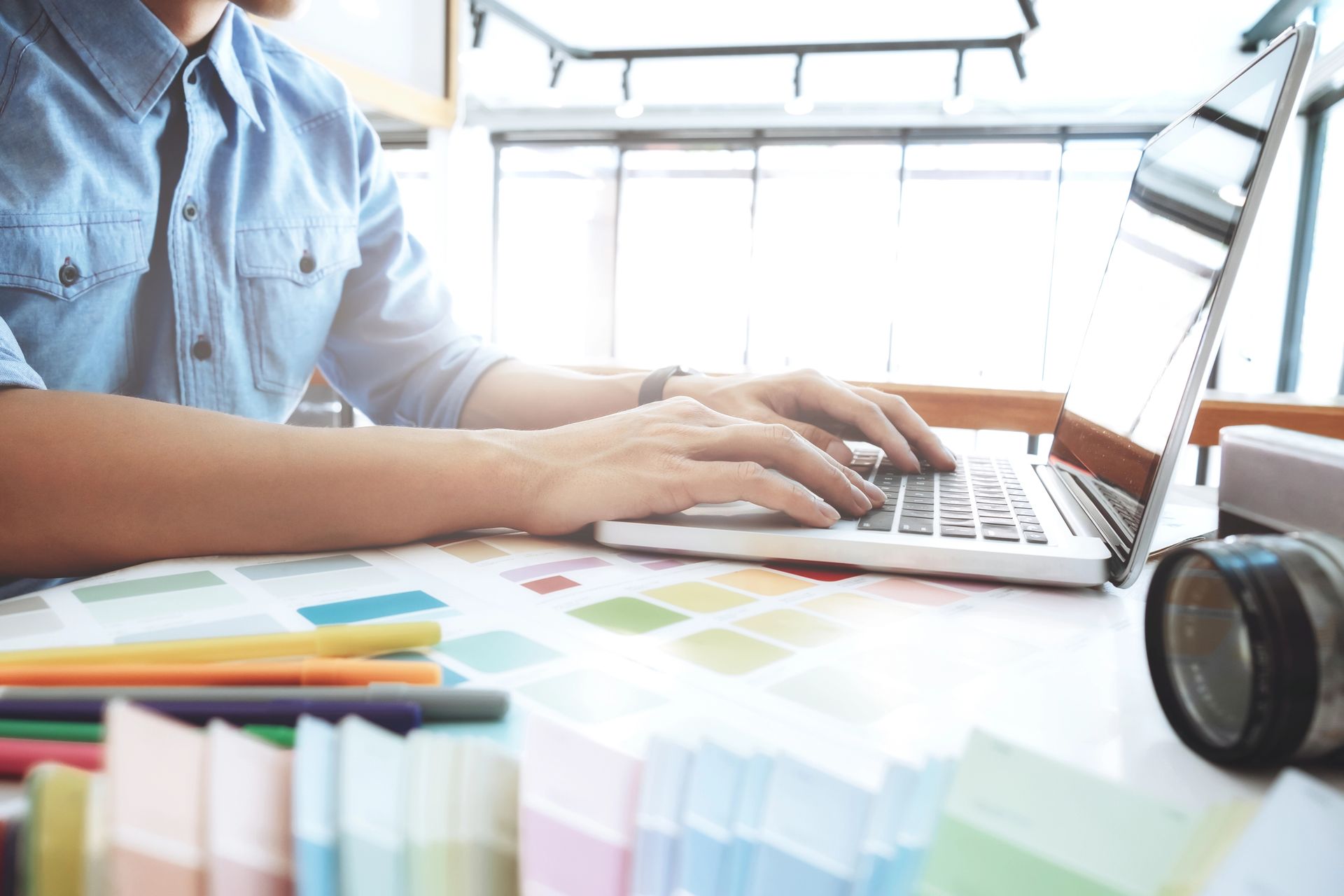 Person typing on a laptop at a desk with colorful charts and a camera lens nearby