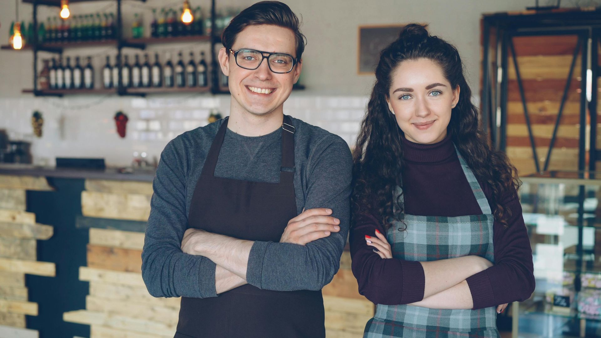 Two smiling café staff in aprons stand with arms crossed behind a counter.