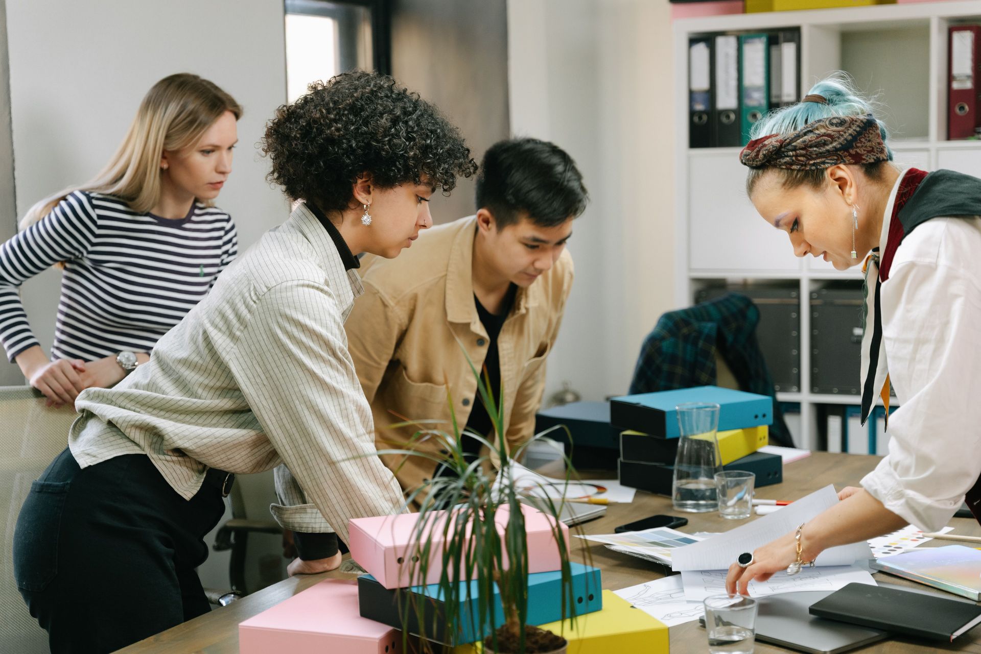 Group of coworkers leaning over a table, reviewing documents and a laptop in a bright office.