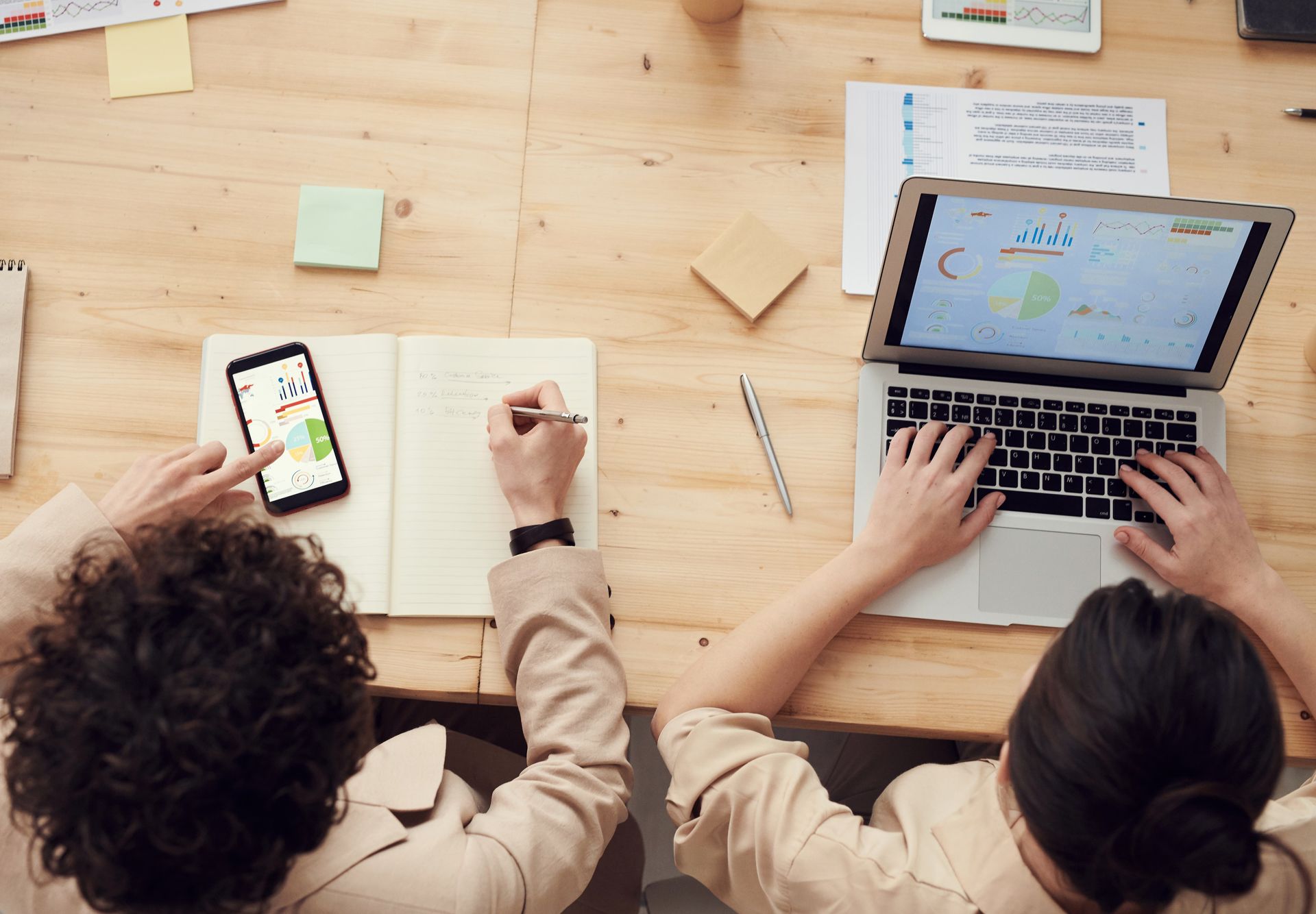 Two people collaborate at a wooden desk, using a laptop, smartphone, and notebook in a bright workspace.