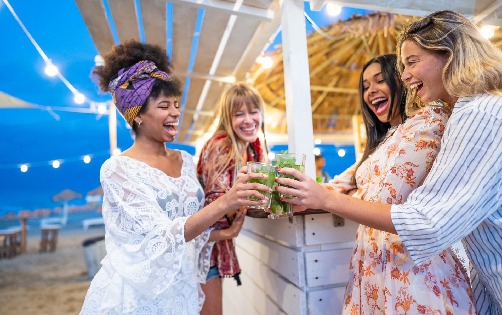A group of women are toasting with drinks at a beach bar.