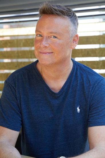 Man in a blue V-neck shirt smiling in front of a window with horizontal blinds