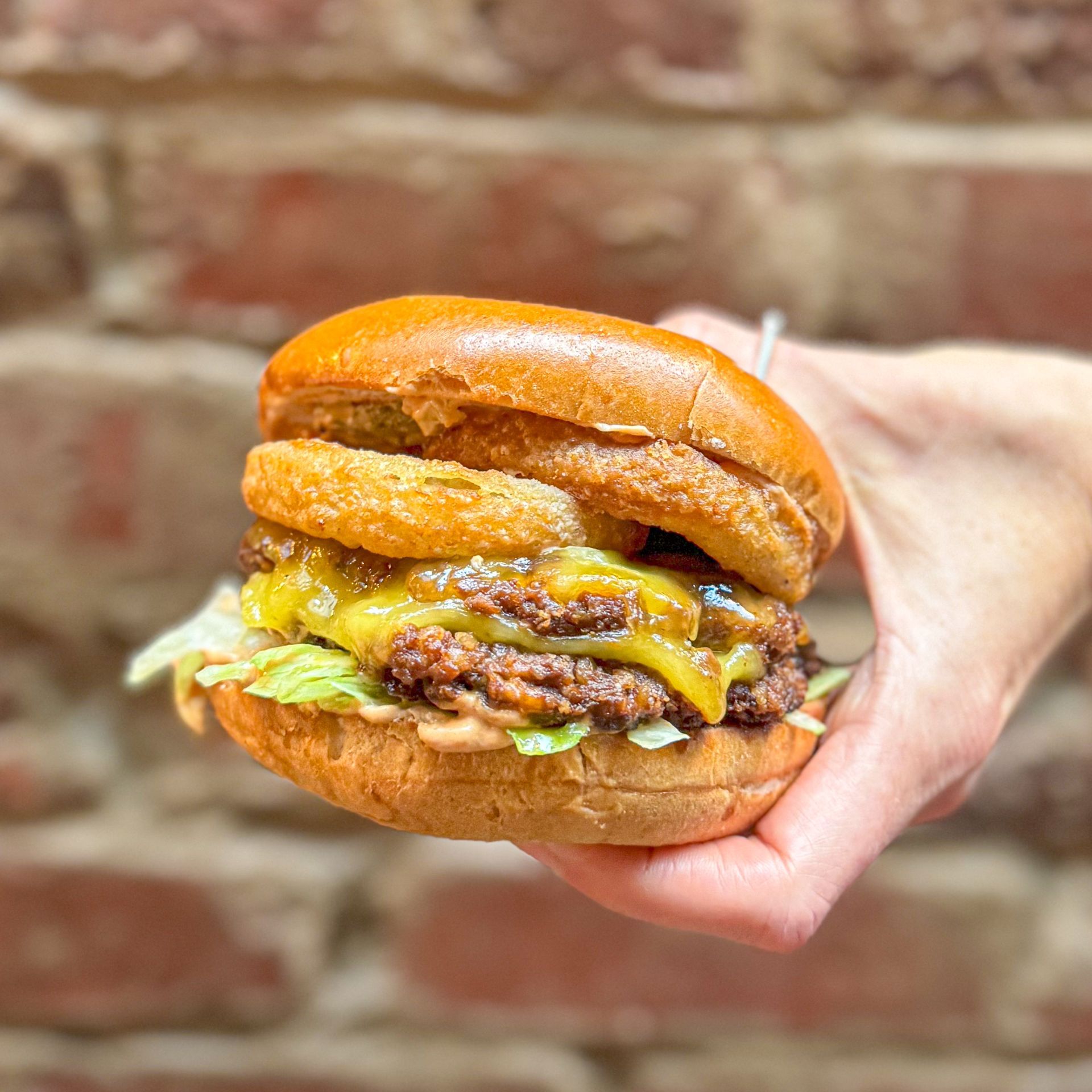 A person is holding a hamburger in their hand in front of a brick wall.