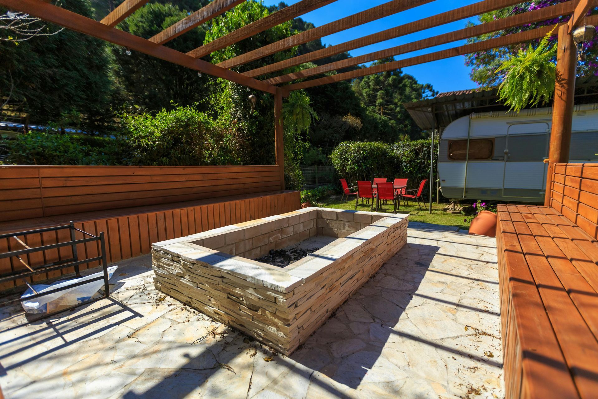 Un foyer en pierre entouré de bancs en bois intégrés sous une pergola sur une terrasse en pierre, avec une caravane garée à proximité.