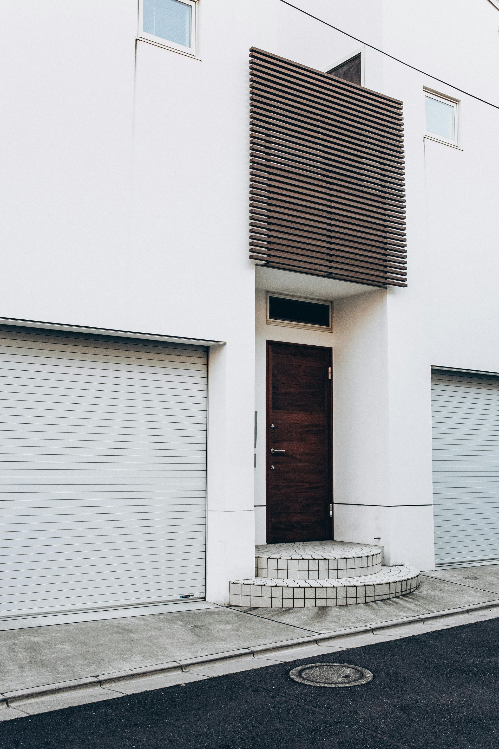 Une maison blanche moderne avec une porte d'entrée en bois foncé, un balcon pergé et deux portes de garage en métal blanc.