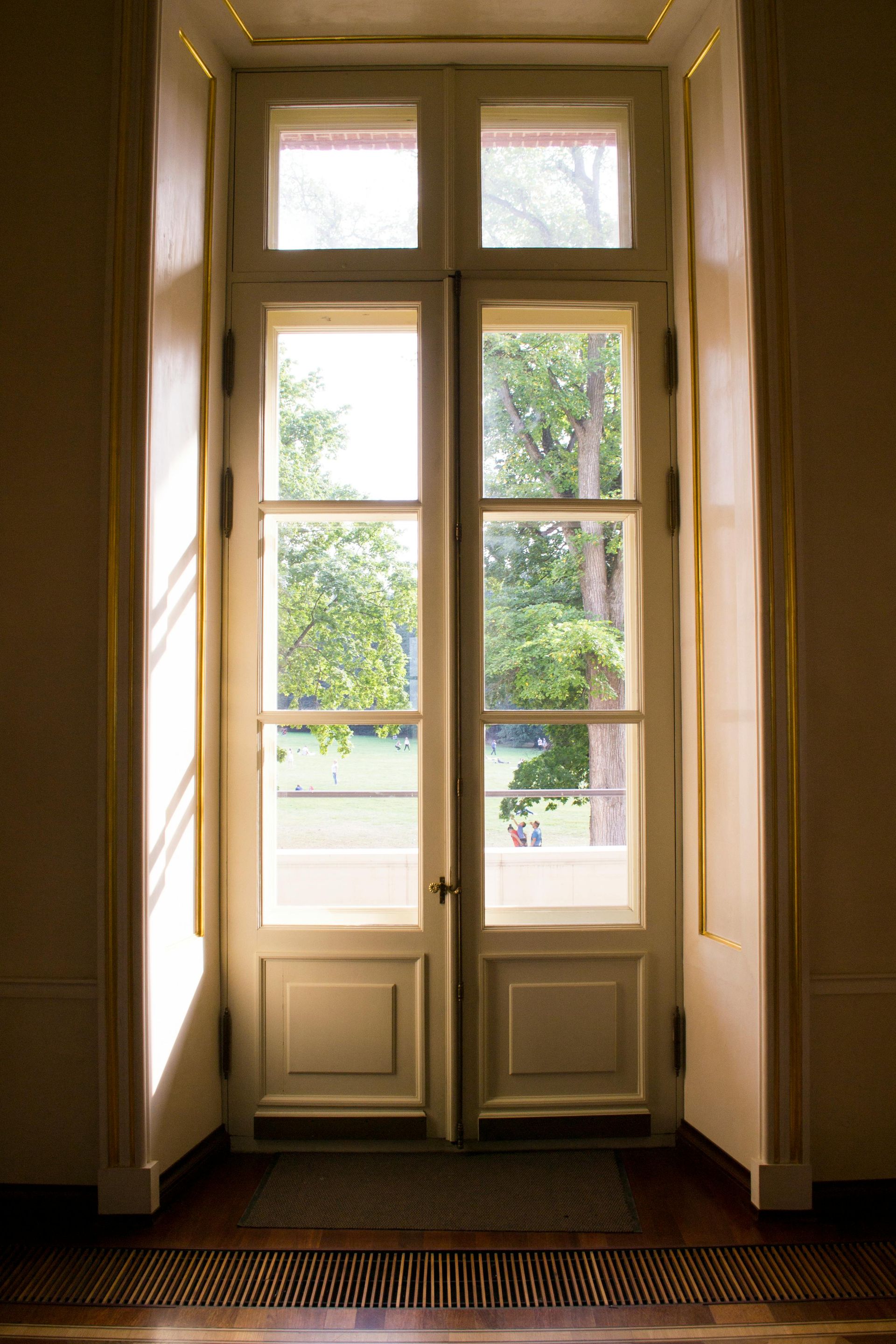 Une haute porte double couleur crème, aux panneaux de verre, donne sur un parc ensoleillé et arboré.