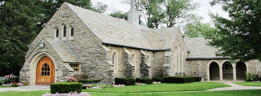 A small stone church with a wooden door is surrounded by trees and grass.
