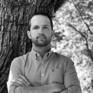 A man with a beard is standing next to a tree with his arms crossed.