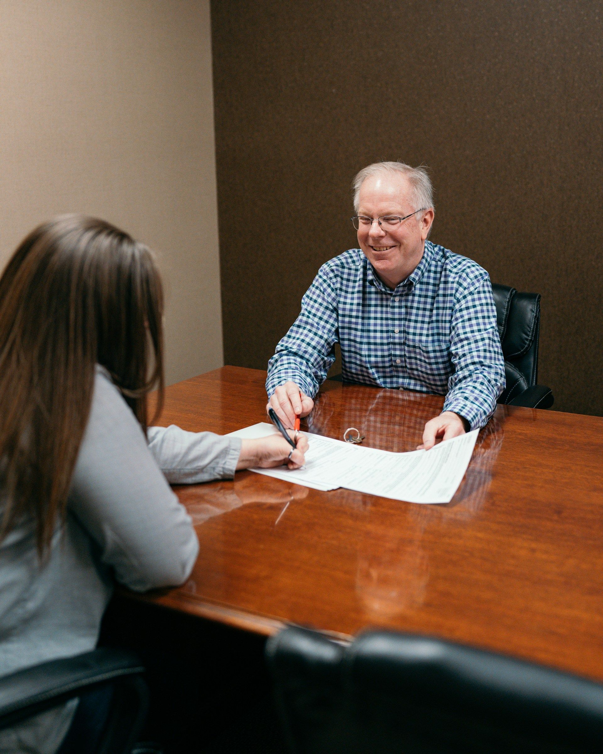 Woman signing document at a table across from a smiling person in a blue shirt.