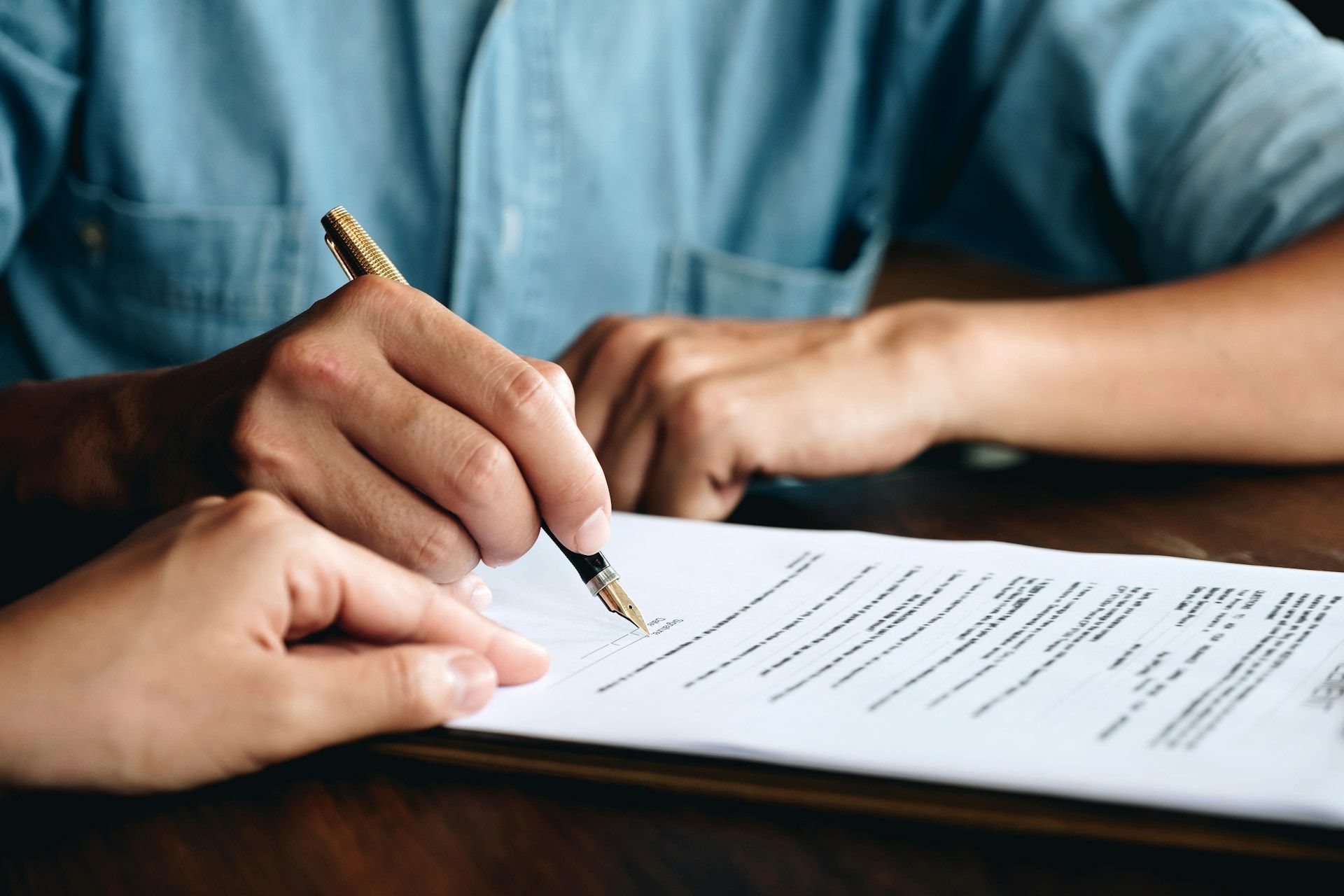 Person signing a document with a pen, another person pointing to a line on the paper.