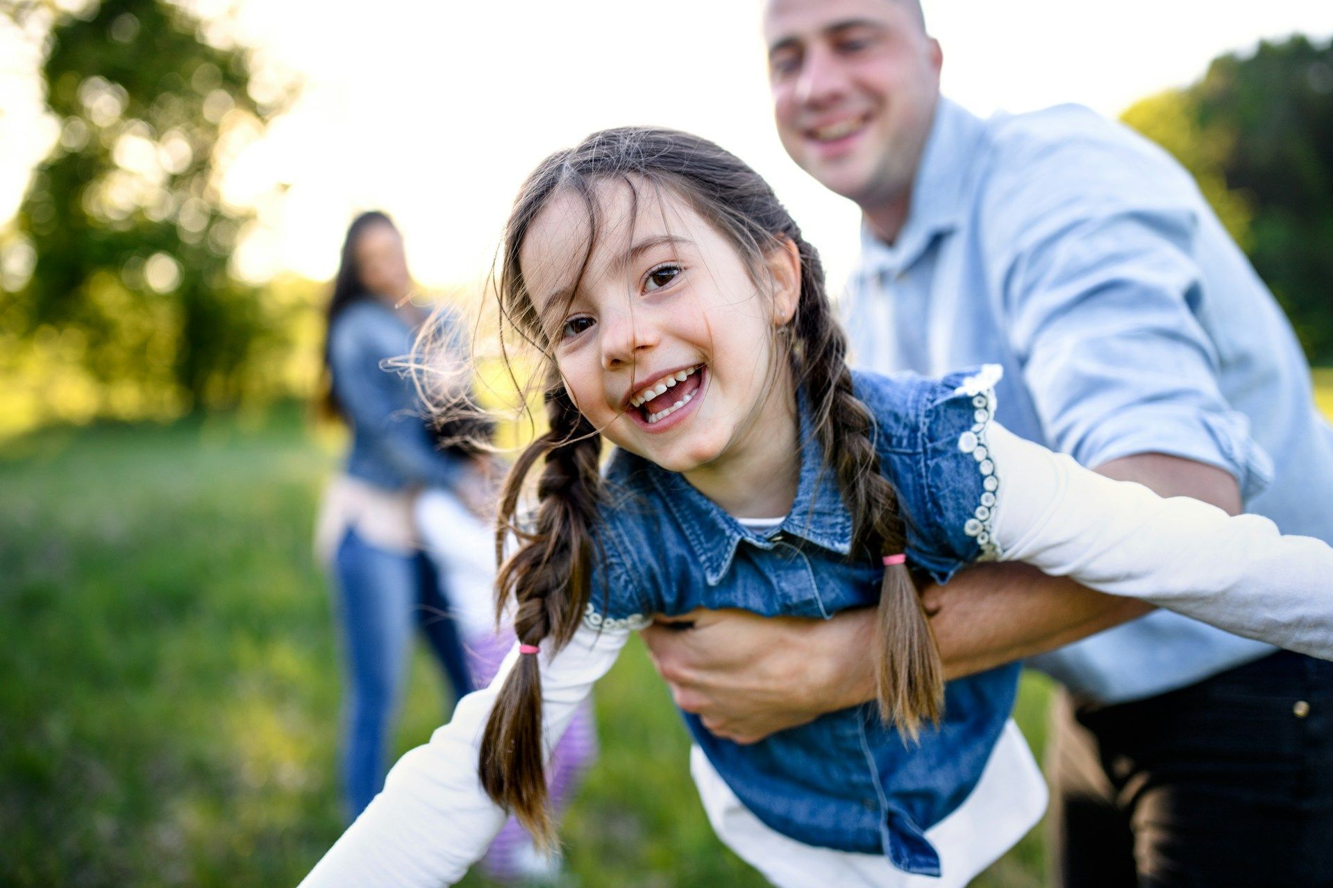Girl smiling, being playfully swung by a man outdoors with a woman in the background.