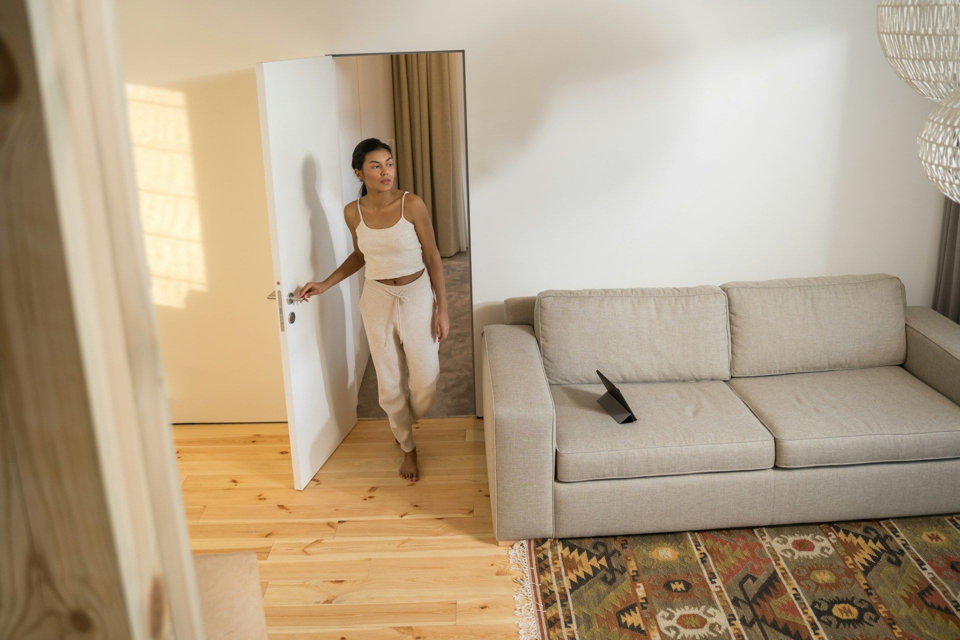 Woman entering a living room, wearing a white outfit. There's a sofa and a rug. Sunlight shines through the doorway.