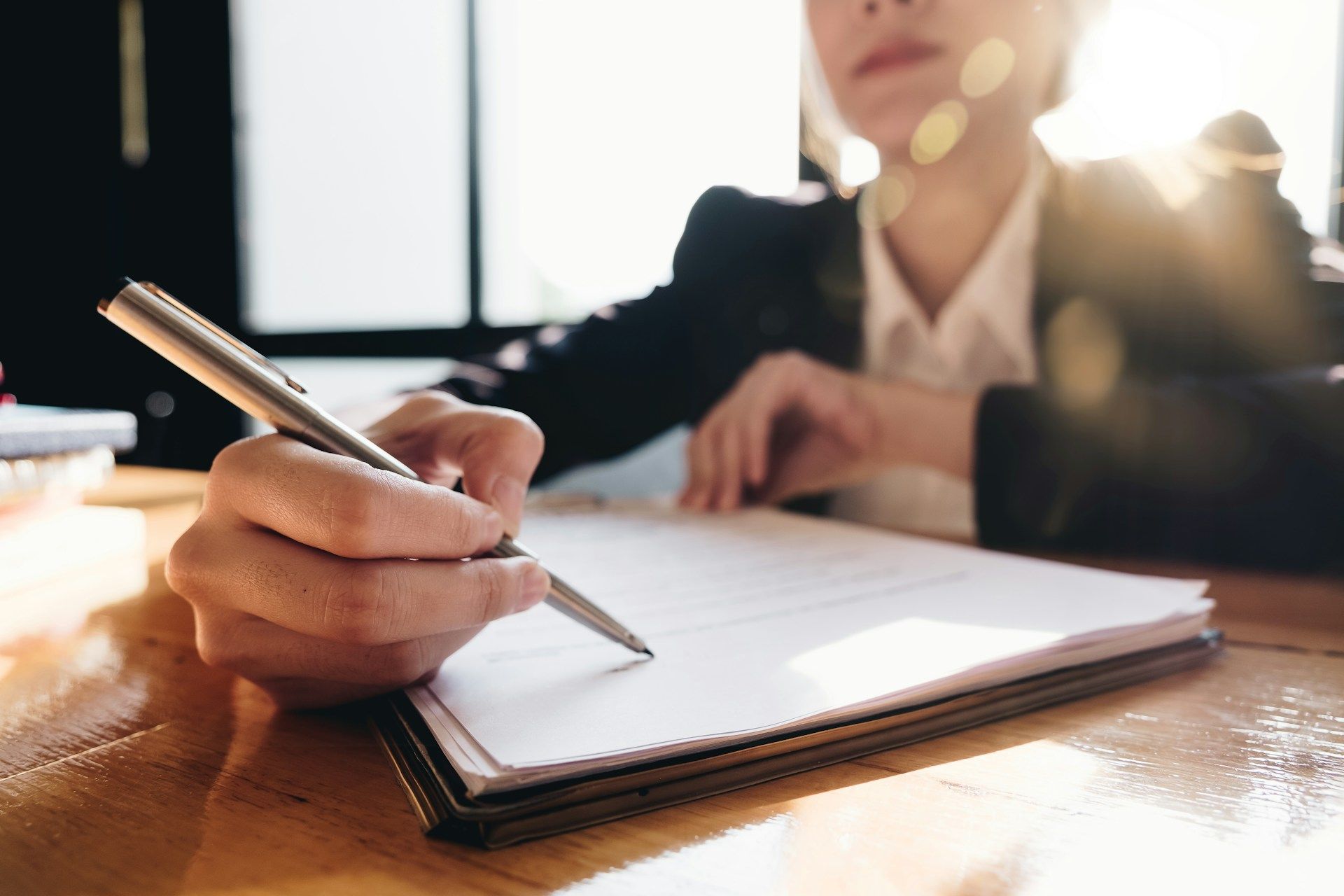 Person in suit writing on a document at a desk, sunlight in background.