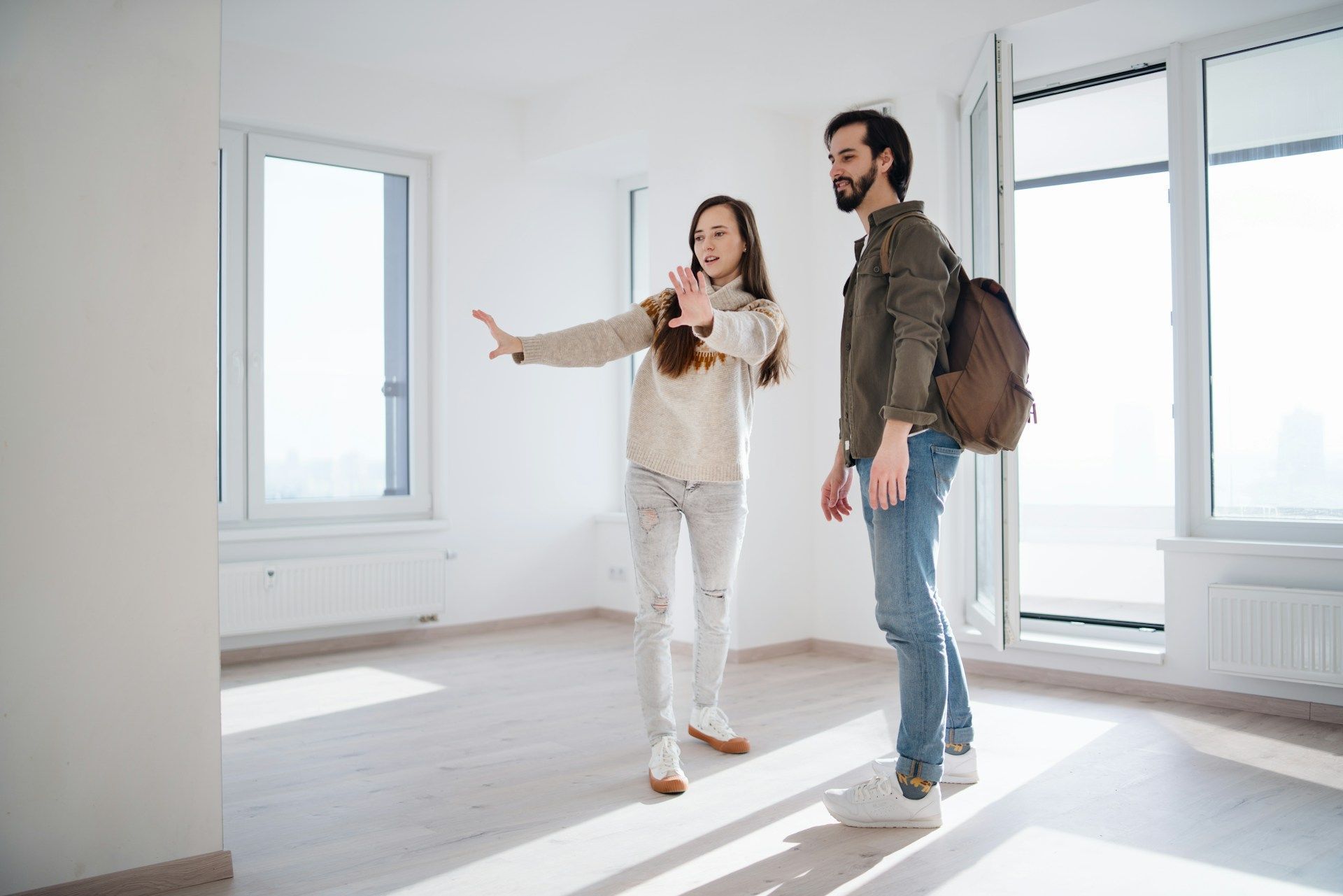 Couple in empty apartment, woman gesturing, man with backpack, looking at windows.