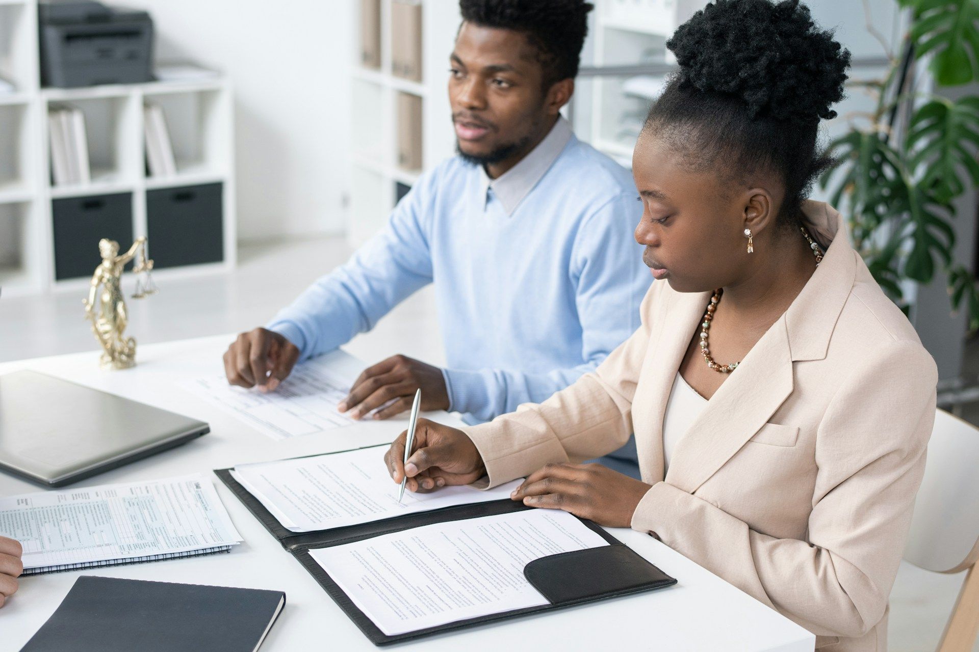 Two people at a desk, reviewing and signing documents. One writes with a pen.